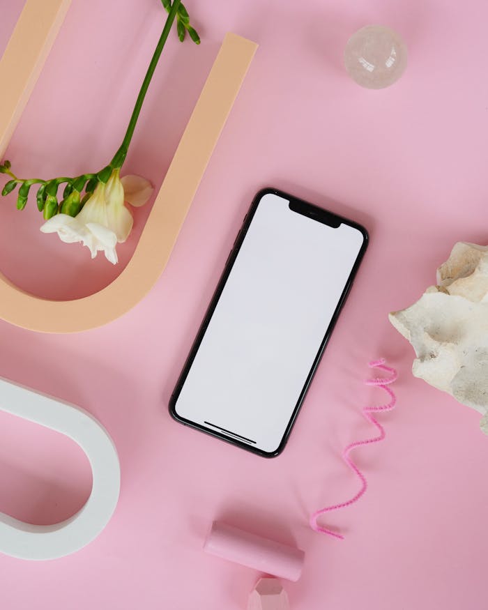 Mockup layout featuring a smartphone, flower, and decorative items on a pink background, ideal for product showcasing.